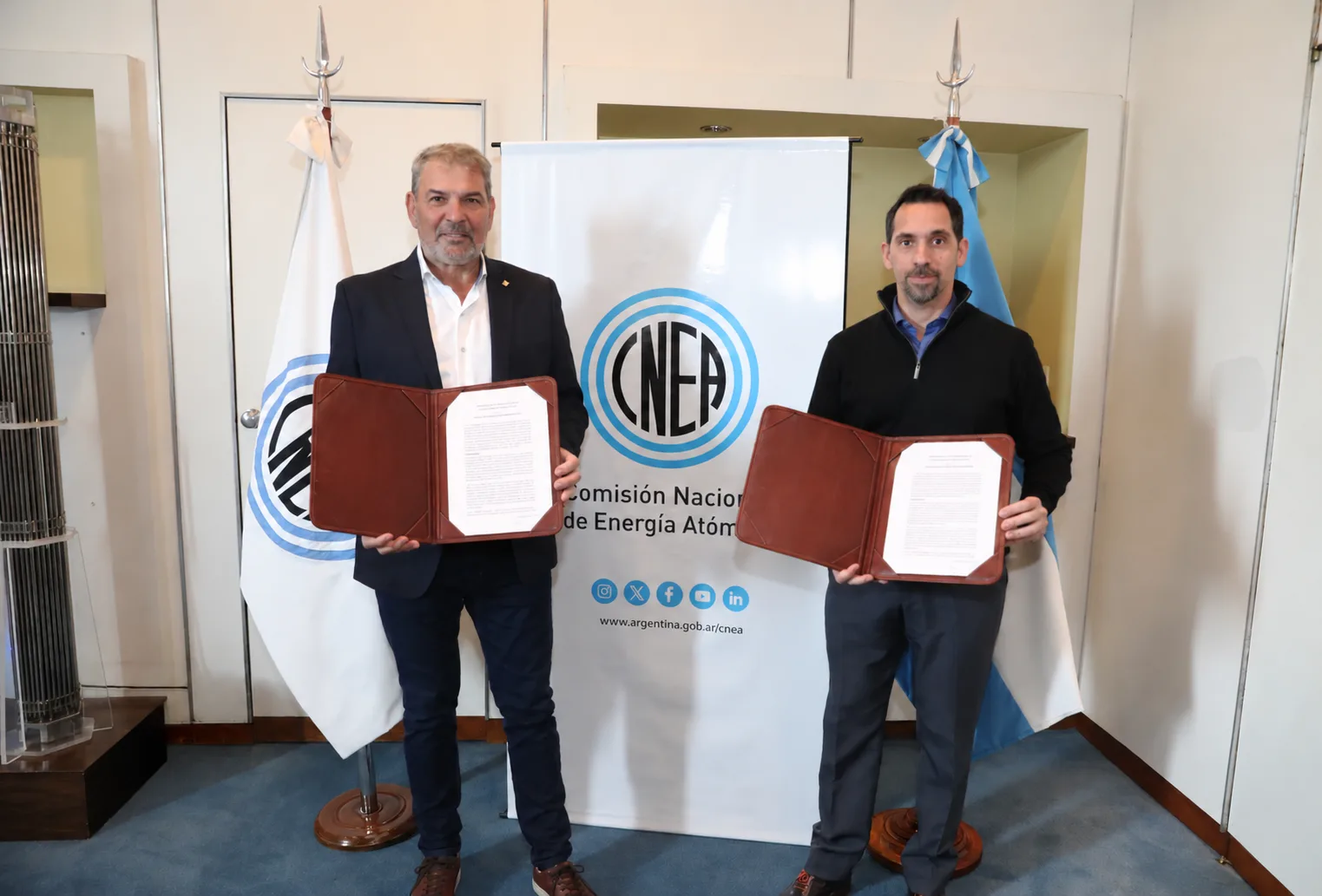 Los presidentes de la AATN y la CNEA muestran los documentos del convenio frente a un banner de la CNEA.